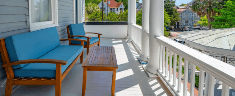 A sunlit historic balcony at 257 Rutledge with grey siding, white columns, and a Haint blue ceiling. The porch is furnished with a wooden outdoor sofa set with bright blue cushions, overlooking a historic neighborhood in Charleston.