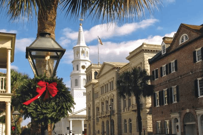 Christmas holiday decorations on a historic street in Charleston, South Carolina, featuring a gas lamp with a green wreath and red bow, with the white steeple of St. Michael's Church in the background.