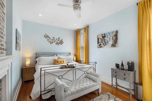 Cozy bed and breakfast guest room featuring light blue walls, a black iron frame bed with white bedding, vibrant yellow curtains framing the windows, and a tufted grey bench at the foot of the bed.