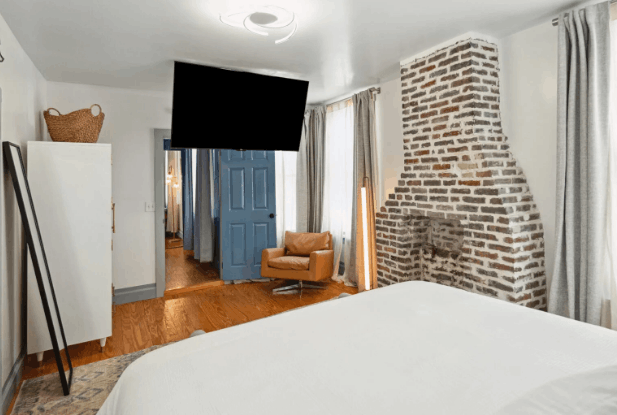 Historic B&B bedroom with exposed brick fireplace, hardwood floors, plush white bed, and leather accent chair.