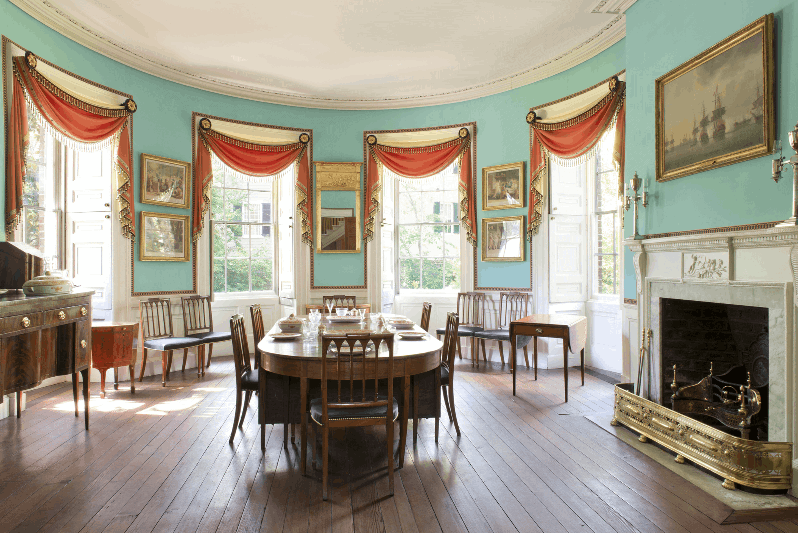 Historic oval dining room inside the Nathaniel Russell House in Charleston, featuring light blue walls, a wooden
  dining table, large draped windows, and a marble fireplace.