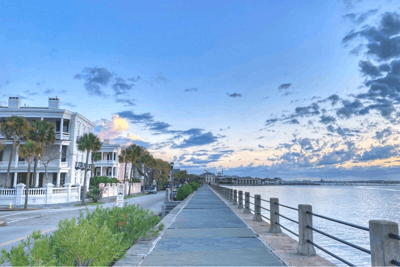 Paved coastal walkway at The Battery in Charleston, South Carolina, lined with historic mansions, palm trees, and the ocean under a sunset sky.