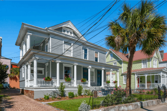 Street view of the historic gray house at 257 Rutledge Inns, featuring classic Southern double-decker porches, white pillars, and a palm tree out front.