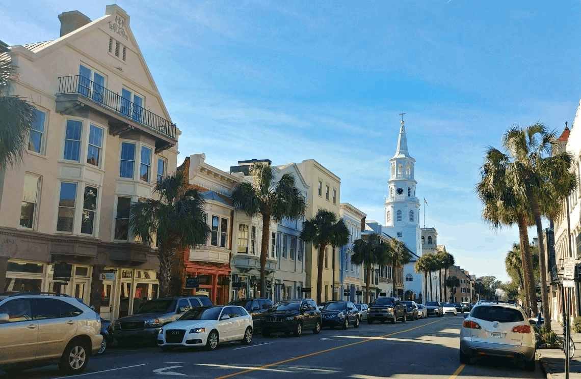 Historic Broad Street in Charleston, South Carolina, featuring colorful multi-story buildings, palm trees, parked
  cars, and the white steeple of St. Michael's Church under a clear blue sky