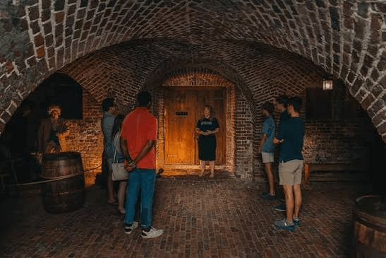 A tour guide addressing a small group of visitors inside a historic brick vault with arched ceilings, featuring a large wooden door and a wooden barrel in the foreground.