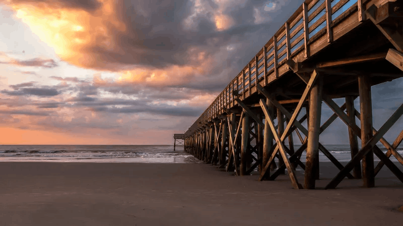 Wooden fishing pier on Isle of Palms beach near Charleston, South Carolina, under a dramatic sunset sky with golden clouds
